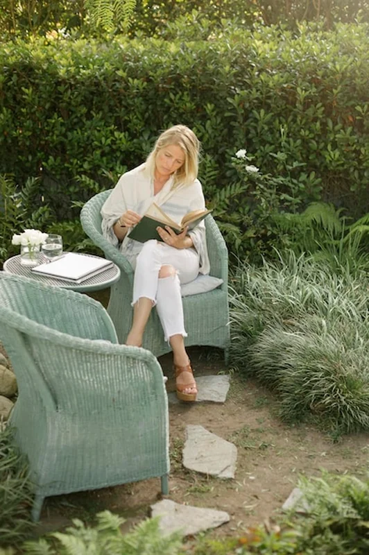 Blond kobieta siedzi na wiklinowym krześle w ogrodzie, czytając